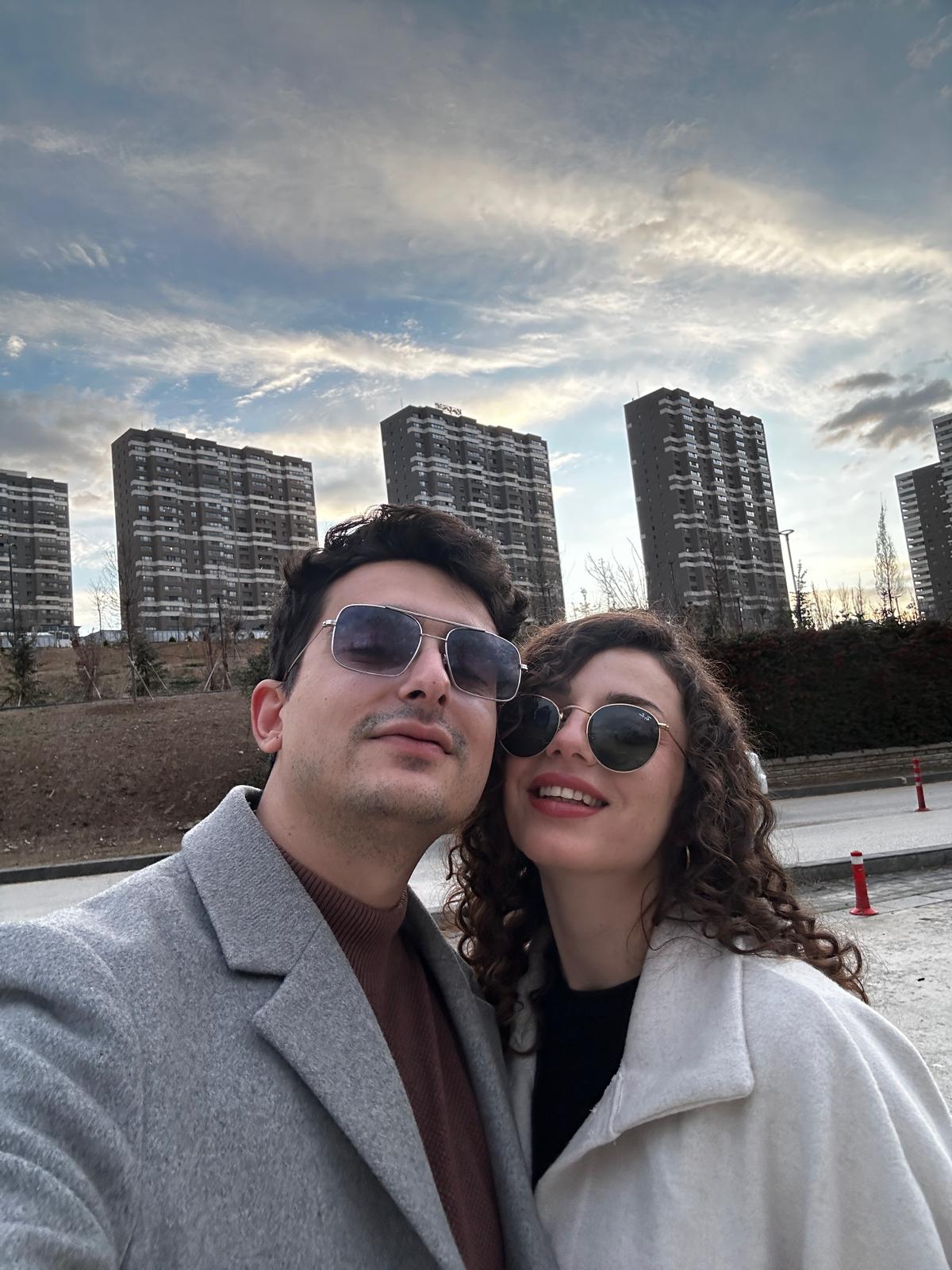 A couple taking a cheerful outdoor selfie in front of modern high-rise apartment buildings under a dramatic cloudy sky.